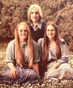 In this vintage formal portrait, three smiling siblings in their 20s pose seated on a grassy field covered in wildflowers. In front is a brunette woman and a blonde woman, both with long hair and eyeglasses. The women are wearing soft, light-colored blouses and skirts. Behind them kneels a blonde man wearing a dark v-necked sweater over a dress shirt.