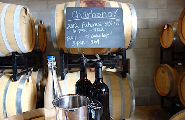 In this winery photo, two bottles of red wine and a glass bottle of water sit next to a steel bucket. Behind them are wooden barrels stacked on racks. A chalk sign advertises wine "futures" available for purchase.