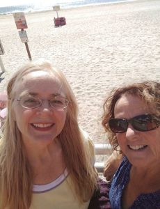 Two women pose against a white beach railing. Behind them stretches a sandy beach with a glimpse of the ocean in the far background. The woman on the left has long straight blonde hair and is wearing glasses and a yellow t-shirt. The woman on the right has shorter brunette curly hair, and is wearing sunglasses and a blue patterned sleeveless top.