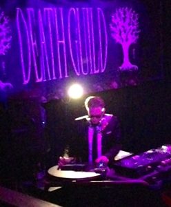 A blonde man in a dark jacket and tie deejays in a night club from a large DJ booth. Purple ambient lighting can be seen over the DJ equipment, and a large banner over his head displays the club logo: the words Death Guild with illustrations of large oak trees on either side.