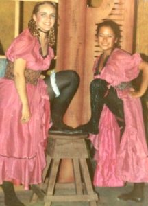 Two young women are dressed as saloon girls for a community college play. They are wearing stage makeup, pink dresses, and black tights. The woman are standing on either side of a wooden stool; each has a leg perched on the stool to show off a ruffled garter.