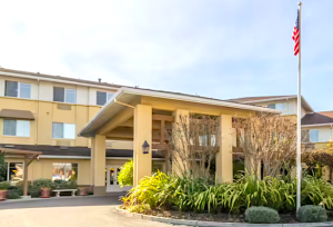 The main entrance of a three-story, yellow residential building for seniors is lined with stoned benches and beautiful landscaping. There is also a tall, shady carport with columns located across from double front doors.