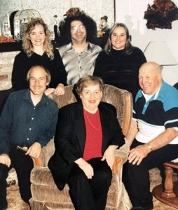 Two senior parents and their adult children pose for a holiday family snapshot circa 1999. In the center of the photo, the mom sits in an upholstered armchair, with her husband seated on the right. Then from left to right behind them are posed their blonde son, blonde daughter, brunette son-in-law, and brunette daughter.