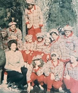 In this vintage snapshot circa 1963, about a dozen family members (adults and children) pose in matching red plaid Christmas pajamas and plaid hats against an outdoor backdrop of California pine trees.