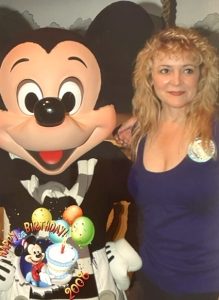 A smiling blonde woman poses next to a costumed Mickey Mouse at Disneyland. She is wearing a navy blue shirt and a birthday pin. On the front of the snapshot is printed a birthday greeting and the year 2008.