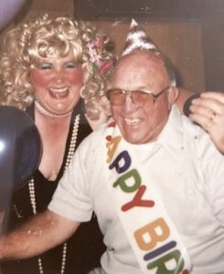 A laughing, 60-year-old man in glasses and a white polo shirt also wears a pointy cardboard birthday hat, a ribbon sash that says "Happy Birthday" in alternating colored letters, and lipstick kisses on his head. Next to him is a laughing, costumed woman in a blonde curly wig, a black dress, and long beaded necklaces that look like pearls.