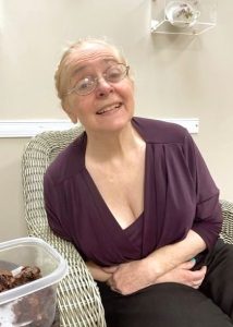 A blonde woman in a purple shirt and eyeglasses is seated in a grey wicker chair. She is leaning all the way over to her right. On the table beside her is a container of homemade brownies.