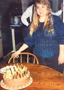 In this vintage snapshot, a 30-year-old woman with long blonde hair, wearing a blue sweater and black pants, stands behind a wooden dining table on which is set a frosted birthday cake ablaze with lit candles.