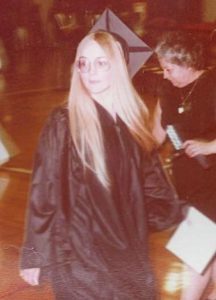 In this vintage snapshot, a young woman dressed in a black graduation cap and gown walks past holding her new junior college diploma. She has long straight blonde hair and is wearing eye glasses.