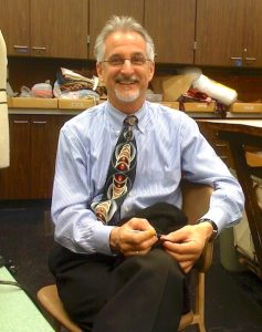A smiling, silver-haired man is seated on a wooden chair in his place of work, a theater's costume shop. He is wearing eye glasses, a blue-striped dress shirt, black dress pants, and a colorful patterned neck tie. On the left of the photo, the edge of a dressmaker's soft mannequin can be glimpsed; in the background can be seen piles of folded fabric and sewing notions stacked on a long counter.