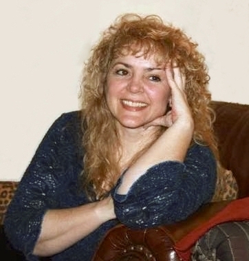 In this snapshot, a blonde woman wearing a dark teal sweater smiles at someone off camera. She is sitting in a dark brown leather chair.