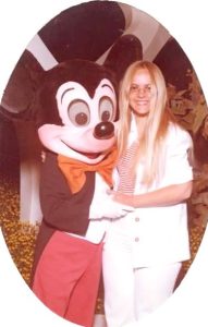 In this vintage snapshot, a smiling blonde teenager, wearing eye glasses and a white summer pantsuit, poses with Mickey Mouse.
