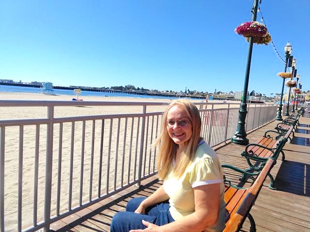 A smiling blonde woman, wearing glasses, a yellow t-shirt and jeans, poses on a wooden bench on the boardwalk. Off to her left, behind a open railing, stretches a sandy beach with a glimpse of the ocean in the far background and a blue sky above.
