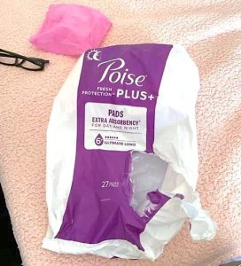 An open package of Poise Plus incontinence pads is set on a peach bedspread next to a pair of eyeglasses.