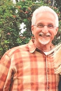 A smiling white-haired man, wearing eye glasses and a red/orange plaid shirt, poses for the camera against a backdrop of greenery.