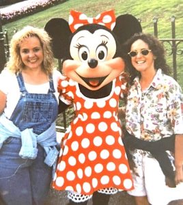 Two smiling women pose with the costumed character Minnie Mouse on the grounds of Disneyland. Minnie wears her traditional red-and-white polka dot dress and bow. On her right is a blonde wearing a white t-shirt and denim overalls and on her left is a brunette wearing sunglasses, a floral button-down shirt, and white shorts.
