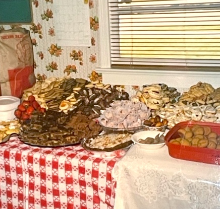 In this vintage snapshot, hundreds of homemade cookies of many different varieties are piled high on platters and dishes that are spread out on two adjacent tables. The tables are covered in festive tablecloths, one checkered red-and-white, the other a silky ivory-on-ivory floral.
