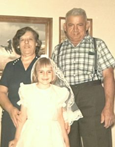 In this vintage snapshot, a little blonde girl, dressed in her first holy communion dress, poses with her grandparents.