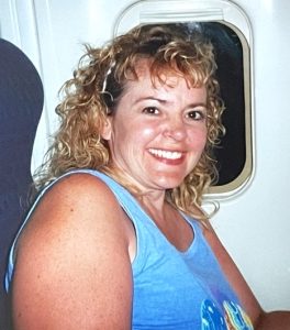 In this modern snapshot, a smiling blonde woman in a sky blue tank top poses in her airplane seat. The shade of the tiny window behind her is open, revealing it is nighttime.