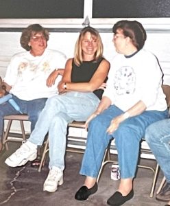 In this vintage snapshot, three women sit on folding chairs during a casual family party. All three are wearing jeans.